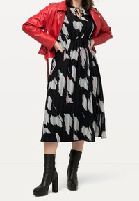 Schwarzes Kleid mit abstraktem weißen und pastellfarbenem Muster, tailliert, kombiniert mit einer leuchtend roten Lederjacke und schwarzen Stiefeln mit hohen Absätzen.