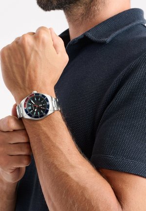 Montre en acier inoxydable argenté avec un cadran noir, des accents rouge et bleu, des repères horaires audacieux, et un bracelet en métal, portée sur un bras bronzé.