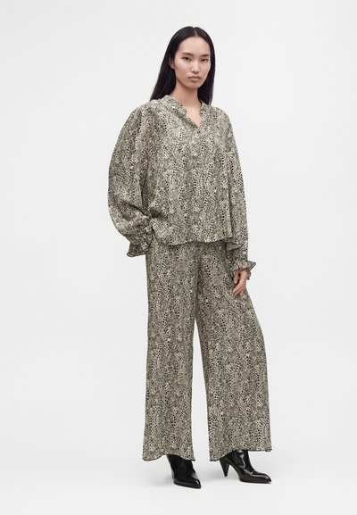 Femme aux longs cheveux raides portant un chemisier ample à motifs et un pantalon large, assortis à des bottes noires à talons, debout devant un fond uni.