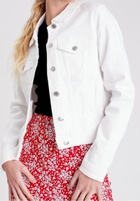 Weiße Jeansjacke mit silbernen Knöpfen, zwei Brusttaschen und langen Ärmeln, getragen über einem schwarzen Oberteil und einem roten Blumenrock.