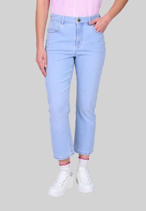 Femme portant un jean court bleu clair, une chemise rayée rose rentrée, des baskets blanches et des chaussettes blanches avec un bord à volants rose, debout devant un fond gris.