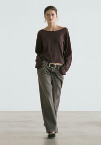 Maglia bordeaux a maniche lunghe con scollo ampio, abbinata a jeans grigi a vita alta. Il modello indossa una cintura nera e scarpe nere, in piedi su una superficie liscia.