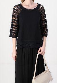 Top nero con maniche in pizzo a tre quarti, orlo in pizzo dettagliato e collo alto, abbinato a una gonna nera fluente e una borsa beige.