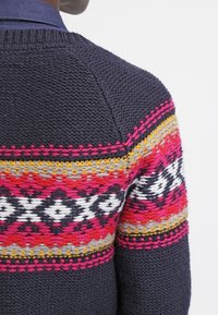 Suéter de punto azul marino oscuro con patrones horizontales multicolores en rosa, rojo, amarillo, blanco y negro en la parte superior de la espalda y las mangas.