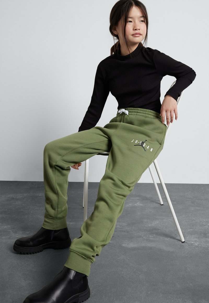 Pantaloni jogger verdi in morbido tessuto con polsini elasticizzati e coulisse bianca. Presenta un logo stampato. Indossati con una maglietta nera a maniche lunghe aderente.