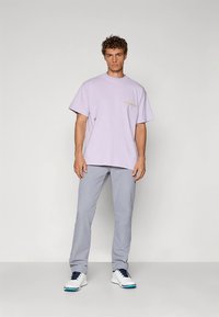 Hellviolettes Baumwoll-T-Shirt mit kurzen Ärmeln und dezentem Schriftzug. Kombiniert mit hellgrauer Hose und weißen Schuhen mit blauen Akzenten.