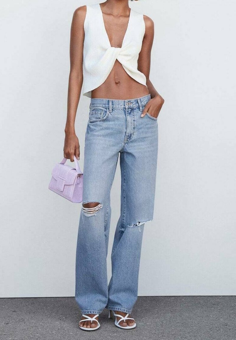 Haut court blanc côtelé avec devant torsadé, jeans larges déchirés bleu clair, et un petit sac à main lavande. Chaussures : sandales blanches.