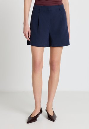 Donna che indossa pantaloncini blu navy su misura, top color bordeaux e décolleté scuri con punta affilata, in piedi su uno sfondo chiaro uniforme.