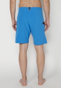 Bañador azul con un color sólido, cinturilla elástica que presenta una etiqueta de marca negra y un pequeño bolsillo lateral. Tejido liso y ligero.