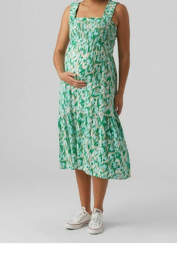 Robe de maternité sans manches, verte à motifs, avec un corsage smocké et un ourlet à volants. Associée à des baskets blanches pour un look décontracté.