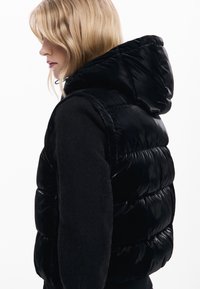 Smanicato imbottito nero con finitura lucida, dotato di cappuccio, design trapuntato e silhouette aderente. Abbinato a una maglia nera a maniche lunghe.