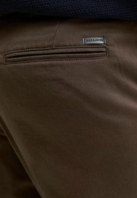 Pantalones marrones con una textura suave, que presentan un bolsillo trasero y una etiqueta "JACK & JONES" cosida en el lado derecho.