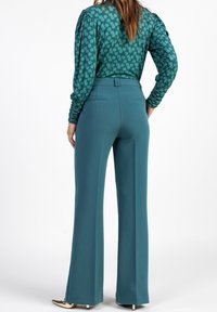 Pantaloni ampi color turchese con una texture liscia, caratterizzati da una vita aderente e due tasche posteriori, abbinati a una blusa verde a fantasia coordinata.