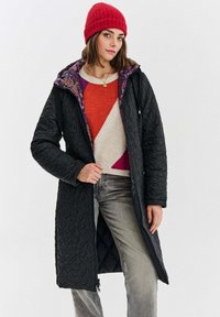 Žena v červené pletené čepici, geometricky barevně rozčleněném svetru, prošívaném černém dlouhém kabátu s květinovou podšívkou a šedých džínech na bílem pozadí.