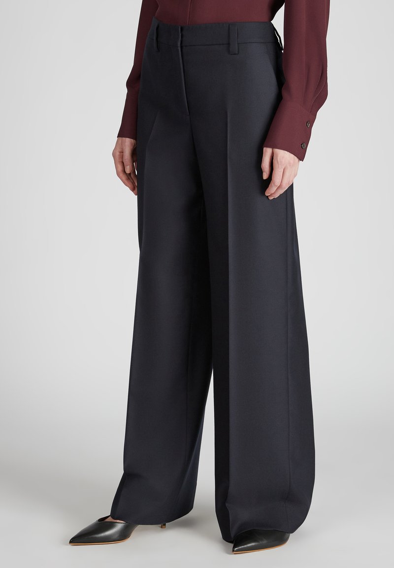 Pantalons larges en tissu bleu marine foncé avec une texture lisse. Dotés d'une taille ajustée et de poches latérales. Associés à des escarpins noirs à bout pointu.