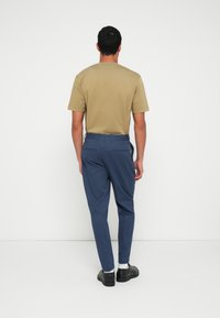 Homem em pé de frente para uma parede branca, vestindo uma camisa de mangas curtas cor de bege, calças azuis, meias brancas e sapatos sociais pretos.