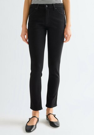 Kobieta w czarnych obcisłych dżinsach, w paskowaną bluzkę włożoną do spodni i czarne buty typu Mary Jane stoi na tle jednolitego jasnego tła.