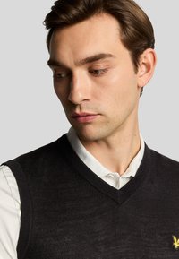 Gilet senza maniche nero con scollatura a V, realizzato in materiale fine, indossato sopra una camicia bianca con colletto, dotata di un piccolo emblema giallo.