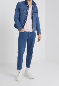 Giacca di jeans dal taglio classico, azzurra con bottoni argentati, indossata sopra una maglietta rosa chiaro con scollo a V e jeans blu aderenti, abbinata a sneakers bianche.