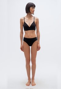 Γυναίκα με κοντά σκούρα μαλλιά, στέκεται ξυπόλυτη φορώντας μαύρο σουτιέν και εσώρουχο, κοιτάζοντας προς τα δεξιά της, μπροστά σε απλό φωτεινό φόντο.