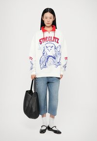 Felpa bianca con dettagli rossi e grafica blu di un gufo, abbinata a jeans cropped blu chiaro. Una borsa nera e scarpe nere completano il look.