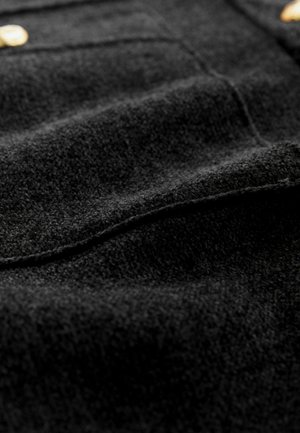 Svart ylletyg med mjuk textur, diskret glans och synliga sömmar; inkluderar detaljer i guldton.