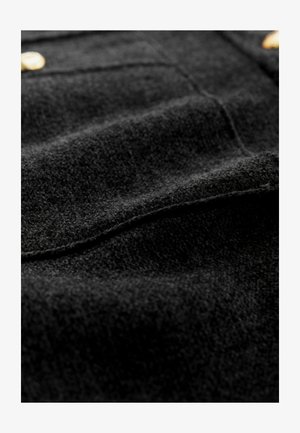 Svart ylletyg med mjuk textur, diskret glans och synliga sömmar; inkluderar detaljer i guldton.