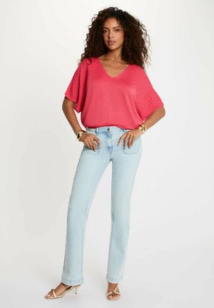 Donna con capelli ricci in posa con le mani sui fianchi, indossa una maglietta rosa a scollo a V, jeans azzurri, braccialetti dorati e tacchi beige.