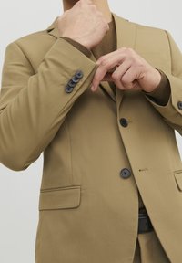 Beige kavaj i strukturerat tyg, med svarta knappar, en slagkrage och en ensam bröstficka, med en brun tröja under.