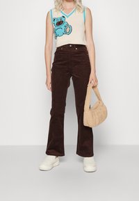 Débardeur en tricot sans manches avec un motif d'ours bleu, pantalon en velours côtelé évasé beige, sac à main texturé beige et chaussures blanches en peluche.