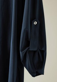 Camisa de manga larga azul marino con tejido texturizado. El puño presenta un botón y un pequeño pliegue, lo que realza el diseño general.