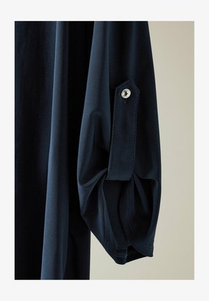 Marineblauwe overhemd met lange mouwen en een gestructureerde stof. De manchet heeft een knoop en een kleine plooitje, wat het algehele ontwerp versterkt.