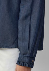 Denim-paita on tummansininen, pehmeästä kankaasta valmistettu, pitkät hihat, joissa on röyhelöetukkeet, sekä kultainen nappikoriste hihassa.