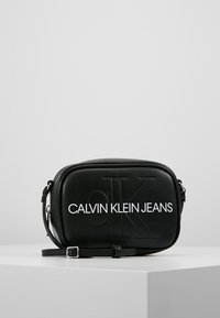 Czarna torba przeszła przez ramię, wykonana z gładkiego materiału syntetycznego. Posiada wytłoczone logo oraz regulowany pasek. Minimalistyczny design z zamkiem błyskawicznym.