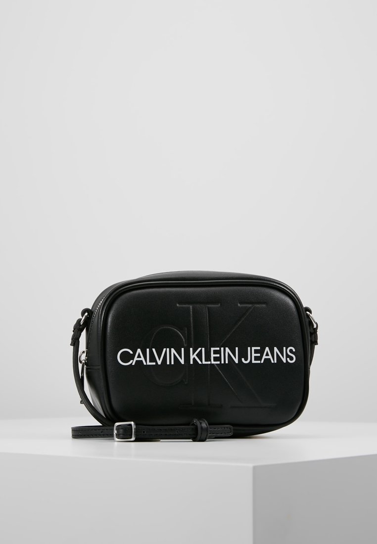 Czarna torba przeszła przez ramię, wykonana z gładkiego materiału syntetycznego. Posiada wytłoczone logo oraz regulowany pasek. Minimalistyczny design z zamkiem błyskawicznym.