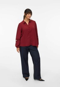 Bordeaux knoop blouse met kraag, pofmouwen en kantdetails bij de manchetten, gecombineerd met donkerblauwe wijde jeans en zwarte hakken.