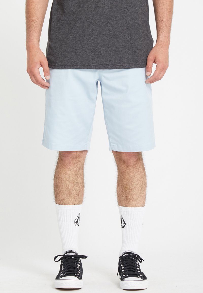 Light blue shorts made of soft fabric, knee-length with a relaxed fit. Paired with black sneakers and white socks featuring a logo.