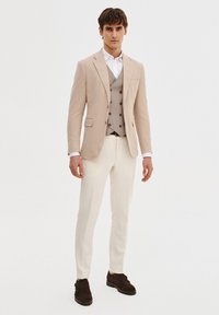 Beżowa lniana marynarka na szarym kamizelce, biała koszula i kremowe spodnie; brązowe zamszowe buty; elegancki design z dopasowanym krojem.