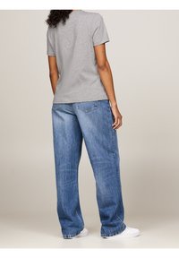 Person iført en grå kortærmet skjorte, løse blå jeans og hvide sneakers, stående med ryggen vendt mod kameraet.