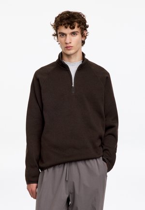 Homme aux cheveux bouclés portant un pull à demi-fermeture éclair marron foncé et un pantalon gris clair, debout devant un fond blanc uni.