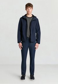 Marineblaue leichte Jacke mit Kapuze, kombiniert mit passenden Slim-Fit-Hosen. Strukturierter grauer Pullover darunter und dunkle Schuhe vervollständigen das Outfit.