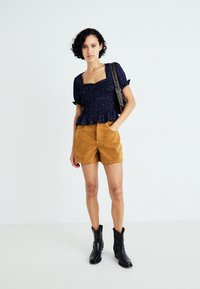 Marineblauwe smocktop met korte pofmouwen, gecombineerd met tan suède shorts en zwarte enkellaarsjes. Model draagt een zwarte tas, minimalistische achtergrond.