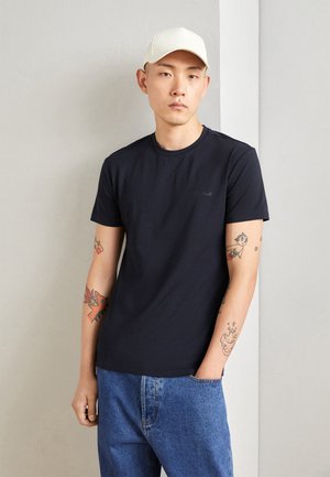 T-shirt bleu marine à manches courtes en coton, avec un col rond et un léger détail de logo. Porté avec un jean en denim bleu.