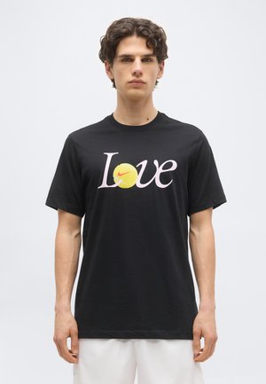 Νεαρός με σγουρά μαλλιά φοράει μαύρο T-shirt με τη λέξη «Love», όπου το γράμμα «o» αντικαθίσταται από μια κίτρινη μπάλα τένις με το κόκκινο σήμα Nike.