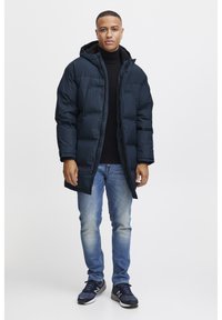 Navy puffer jacket with a hood, zip closure, and wide quilted pattern, worn over a black turtleneck, paired with light blue jeans and sneakers.