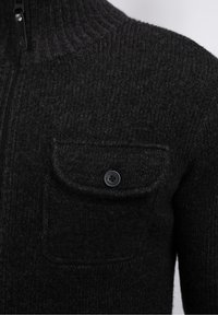 Grå stickad tröja med hög krage, som har dragkedja och en bröstficka med knapp. Texturerad tyg ger djup.