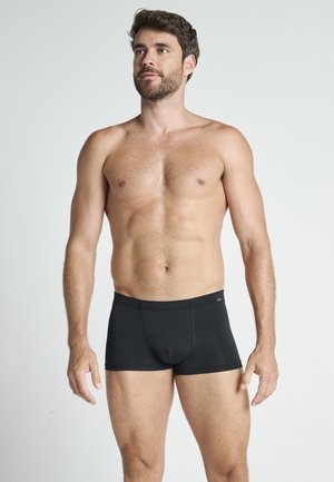 Schwarze Herren-Boxershorts mit enganliegendem Schnitt, weichem Stoff und elastischem Bund, präsentiert an einem Mann, der vor einem neutralen Hintergrund steht.