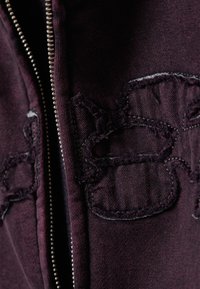 Nærbillede af maroon stof med en metal lynlås og slidte sort syede lapper i abstrakte blomstermønstre.