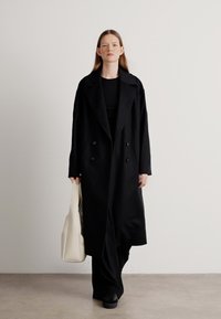 Abrigo negro de doble botonadura con grandes solapas, llevado sobre un conjunto negro, combinado con un bolso tote blanco y zapatos negros.