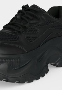 Baskets noires épaisses avec semelle épaisse, tige en mesh et cuir, et lacets ronds sur fond uni.
